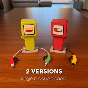 May include: Two charging stations shaped like fuel pumps, one yellow and one red, each with a base and charging cables. The yellow pump features the text POWER PUMP, while the red pump displays a battery icon. Text reads 2 VERSIONS Single & double cable.