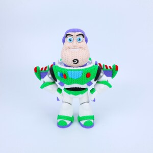 Könnte beinhalten: Eine gehäkelte Figur eines Weltraum-Ranger-Charakters mit violetter Kapuze, weiß-grünem Brustpanzer und blauen Akzenten. Die Figur weist eine strukturierte Strickoberfläche, große blaue Augen und ein kleines schwarzes Detail am Kinn auf.