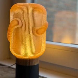 Pode incluir: Uma moderna luminária de mesa laranja com um design cilíndrico. A luminária apresenta uma sombra texturizada e canelada e uma base preta com um detalhe em madeira. A luminária está acesa, emitindo um brilho quente. A luminária tem cerca de 30 cm de altura.