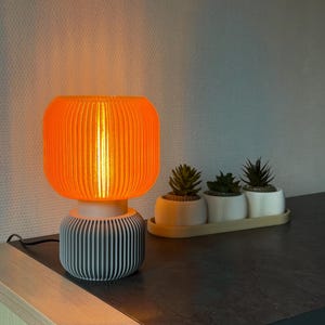 Op de afbeelding: Oranje en grijze tafellamp met een geribbelde kap en voet. De lamp is ingeschakeld en geeft een warme gloed. Drie kleine vetplanten in potten staan op de achtergrond en voegen een vleugje groen toe.