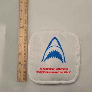 Puede incluir: Bolsa blanca Shark Week Emergency Kit con un gráfico de boca de tiburón azul y texto rojo. La bolsa mide aproximadamente 13 cm de largo, medido con una regla.