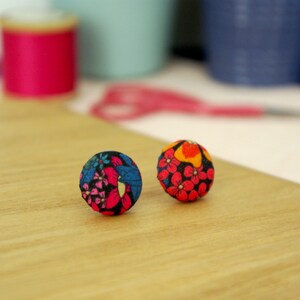 May include: Two fabric-covered ball-shaped earrings with floral patterns. One earring features a blue, pink, and black floral design, while the other has a red, orange, and yellow floral pattern. They are set on a light wooden surface.