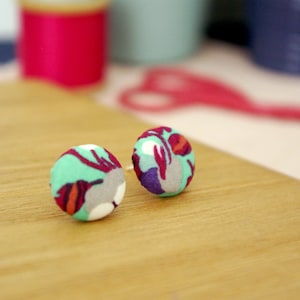 May include: A pair of round stud earrings with a fabric covering. The fabric has a pattern of teal, red, purple, and gray shapes on a white background. The earrings are on a wooden surface.