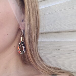 May include: A pair of dangle earrings with a gold accent. The earrings are made of fabric with a floral pattern in shades of orange, blue, and green.