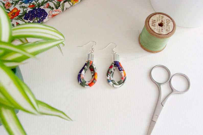 May include: A pair of floral fabric earrings with silver hooks. The earrings are a teardrop shape and are made of white fabric with a colorful floral pattern. The fabric is folded over itself to create a double layer effect.