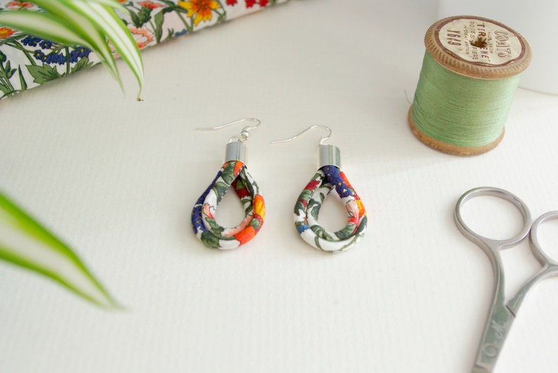 May include: A pair of floral fabric earrings with silver hooks. The fabric is white with a floral pattern in shades of orange, green, blue, and white.