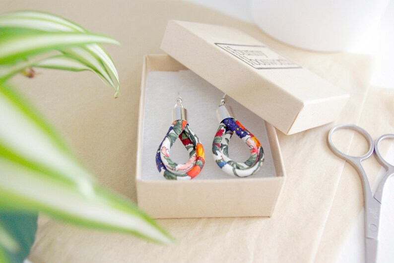 May include: A pair of floral fabric earrings with silver hooks. The earrings are in a white box with a white background. The earrings are a teardrop shape and have a floral pattern in shades of blue, green, orange, and white.