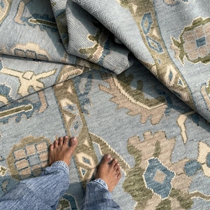 May include: A light blue rug with a detailed pattern in beige, green, and blue. The rug is folded, showing its textured weave. The design incorporates floral and geometric shapes, giving a traditional appearance.