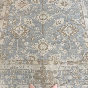 May include: A large area rug featuring a repeating geometric pattern in shades of light blue and beige. The design includes central diamond-shaped motifs and intricate floral-inspired details, finished with a wide, decorative border along the edge.