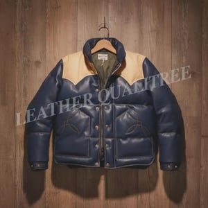 May include: A navy blue and tan leather puffer jacket with a quilted design. The jacket has a high collar and a zip-up front. The text "LEATHER QUALITREE" is visible on the jacket. The jacket is hanging on a wooden hanger.
