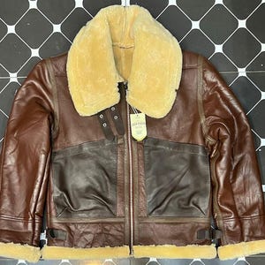 Può includere: Giacca in pelle marrone con colletto e fodera in montone color crema. La giacca presenta una chiusura con zip, due tasche frontali e un dettaglio con cinturino in pelle. Un'etichetta recita "LEATHER SHEEP".