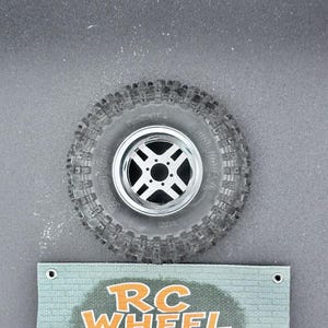 K&ouml;nnte beinhalten: Ein einzelnes RC-Autorad mit einem schwarzen Reifen und einer silbernen Felge. Der Reifen hat ein robustes Profil. Ein Banner unter dem Rad zeigt "RC WHEEL SHOP T" in orangefarbenem und wei&szlig;em Text.