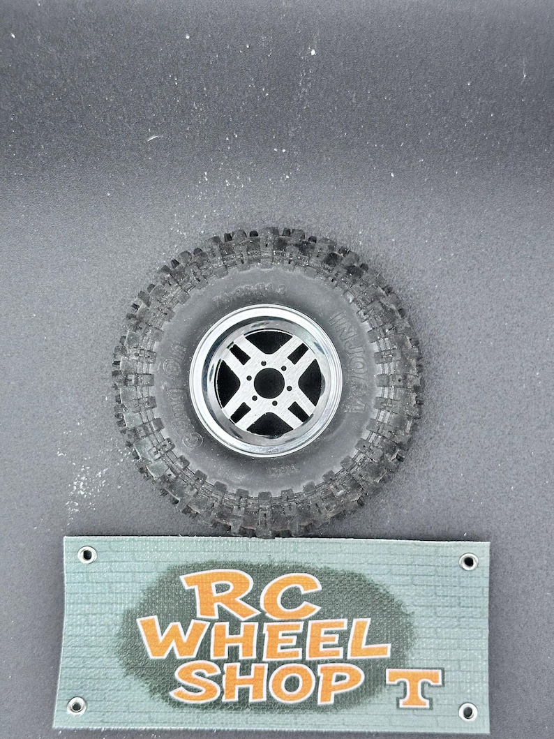 K&ouml;nnte beinhalten: Ein einzelnes RC-Autorad mit einem schwarzen Reifen und einer silbernen Felge. Der Reifen hat ein robustes Profil. Ein Banner unter dem Rad zeigt "RC WHEEL SHOP T" in orangefarbenem und wei&szlig;em Text.