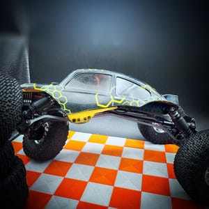 Könnte beinhalten: Ein schwarz-gelbes ferngesteuertes Auto mit einem Wabenmuster auf der Karosserie. Das Auto hat große, grobe Reifen und steht auf einer karierten orange-weißen Oberfläche. Der Hintergrund ist dunkel.