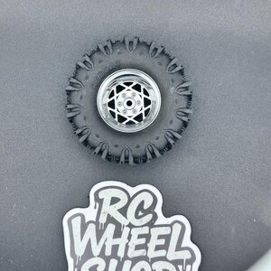 K&ouml;nnte beinhalten: Ein schwarzer RC-Autoreifen mit silberner Felge und Sternmuster. Der Reifen hat ein robustes Profil, und das Wort "RED CAT" ist auf der Seitenwand zu sehen. Ein Aufkleber mit dem Text "RC WHEEL SHOP" ist ebenfalls vorhanden.
