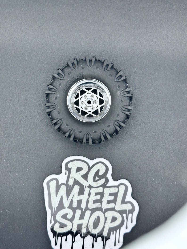 K&ouml;nnte beinhalten: Ein schwarzer RC-Autoreifen mit silberner Felge und Sternmuster. Der Reifen hat ein robustes Profil, und das Wort "RED CAT" ist auf der Seitenwand zu sehen. Ein Aufkleber mit dem Text "RC WHEEL SHOP" ist ebenfalls vorhanden.