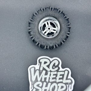 K&ouml;nnte beinhalten: Ein schwarzer RC-Autoreifen mit silberner Felge, mit dem Text "REDCAT" auf dem Reifen. Unter dem Reifen befindet sich ein Aufkleber mit dem Text "RC WHEEL SHOP" in einer tropfenden Schrift.