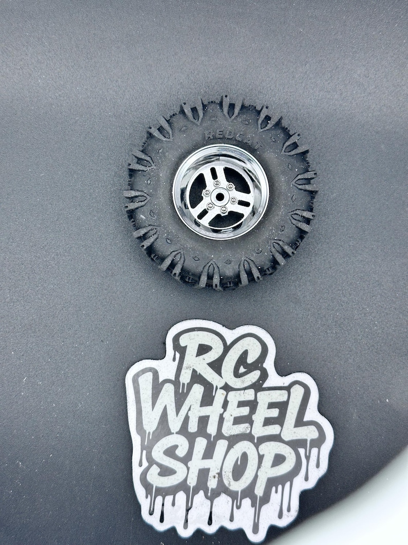 K&ouml;nnte beinhalten: Ein schwarzer RC-Autoreifen mit silberner Felge, mit dem Text "REDCAT" auf dem Reifen. Unter dem Reifen befindet sich ein Aufkleber mit dem Text "RC WHEEL SHOP" in einer tropfenden Schrift.