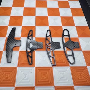 Könnte beinhalten: Vier schwarze Kohlefaserkomponenten mit unterschiedlichen offenen Designs sind auf einer karierten orange-weißen Oberfläche angeordnet. Die Komponenten haben ein strukturiertes Aussehen und sind wahrscheinlich für ein mechanisches Gerät.