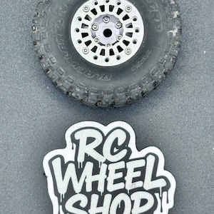 Puede incluir: Primer plano de un neumático negro con una llanta plateada. La llanta tiene múltiples agujeros y está atornillada al neumático. Debajo, una pegatina blanca con las palabras "RC WHEEL SHOP" en una fuente goteante.