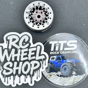 Op de afbeelding: Een zilveren en zwarte RC-wiel met een stervormig ontwerp. De afbeelding bevat ook twee stickers: een met de tekst "RC WHEEL SHOP" en een andere met "TiTS ROCK CRAWLING" en een blauwe RC-truck.
