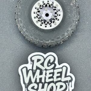 Könnte beinhalten: Nahaufnahme eines schwarzen RC-Autoreifens mit einer silbernen Felge. Die Felge hat ein Sternmuster mit mehreren kleinen Schrauben. Darunter befindet sich ein Aufkleber mit dem Text "RC WHEEL SHOP" in Weiß und Schwarz.