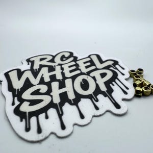 May include: An image featuring a sticker with the text "RC WHEEL SHOP" in a dripping font style, accompanied by a small pile of gold-colored nuts. The sticker is black and white.
