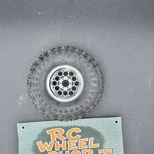 May include: A black RC car tyre with a silver wheel featuring a honeycomb design. The tyre has a textured tread pattern. Below the tyre is a green banner with the text "RC WHEEL SHOP T" in orange.