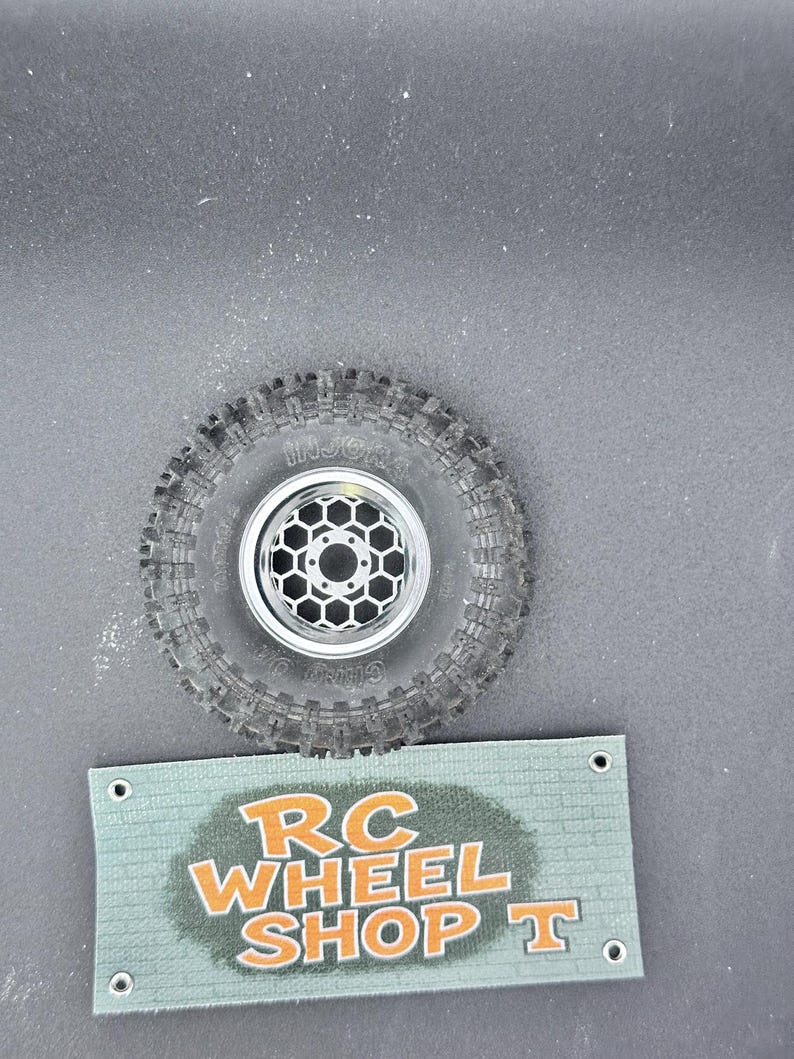 K&ouml;nnte beinhalten: Ein schwarzer RC-Autoreifen mit einer silbernen Felge mit Wabenmuster. Der Reifen hat ein strukturiertes Profil. Darunter befindet sich ein gr&uuml;nes Banner mit dem Text "RC WHEEL SHOP T" in Orange.