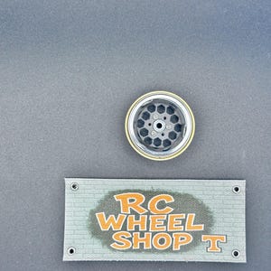 K&ouml;nnte beinhalten: Ein schwarzes und silbernes RC-Autorad mit goldenem Rand. Das Rad hat eine Reihe sechseckiger Aussparungen. Darunter befindet sich ein Banner mit dem Text "RC WHEEL SHOP T" in Orange.