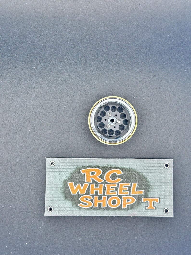K&ouml;nnte beinhalten: Ein schwarzes und silbernes RC-Autorad mit goldenem Rand. Das Rad hat eine Reihe sechseckiger Aussparungen. Darunter befindet sich ein Banner mit dem Text "RC WHEEL SHOP T" in Orange.