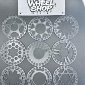 Puede incluir: Se muestran nueve diseños de ruedas RC plateadas, cada uno con un patrón único de agujeros y radios. Encima, una pancarta con el texto "RC WHEEL SHOP" en una fuente de estilo grafiti. Las ruedas están dispuestas en una cuadrícula de 3x3.