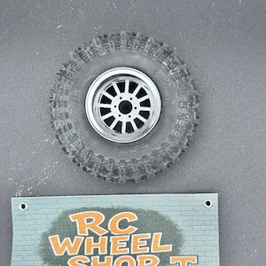 K&ouml;nnte beinhalten: Ein schwarzer RC-Autoreifen mit einer silbernen Felge mit einem Mehrspeichen-Design. Der Reifen hat ein robustes Profil. Darunter befindet sich ein t&uuml;rkisfarbenes Schild mit dem Text "RC WHEEL SHOP T" in Orange.