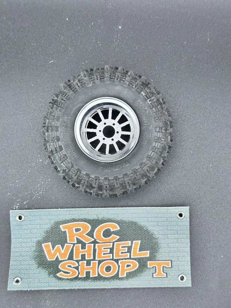 K&ouml;nnte beinhalten: Ein schwarzer RC-Autoreifen mit einer silbernen Felge mit einem Mehrspeichen-Design. Der Reifen hat ein robustes Profil. Darunter befindet sich ein t&uuml;rkisfarbenes Schild mit dem Text "RC WHEEL SHOP T" in Orange.