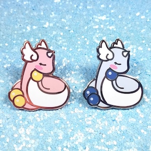 May include: Two enamel pins featuring a cartoon dragon design. The dragon on the left is pink with yellow accents and the dragon on the right is light blue with dark blue accents.