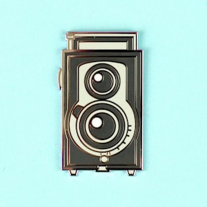 Puede incluir: Un pin de esmalte plateado de una cámara vintage con dos lentes. La cámara es de color negro y blanco con la parte superior de color crema.