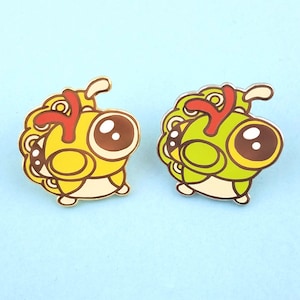 May include: Two enamel pins featuring a cartoon butterfly with large eyes, a red antenna, and a yellow or green body.