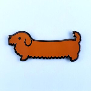 May include: Orange dachshund enamel pin with black outline. The pin is shaped like a dachshund dog with a long body and short legs.