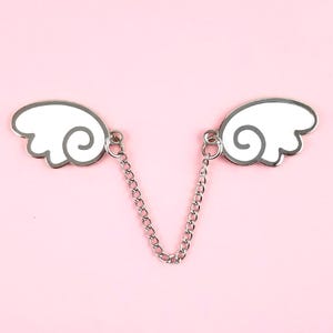 Puede incluir: Una cadena de plata con dos alas de esmalte blanco unidas. Las alas tienen un diseño simple y de dibujos animados.