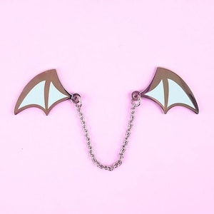 Puede incluir: Un par de alas de metal plateadas con detalles en esmalte blanco están conectadas por una cadena plateada. Las alas tienen forma de alas de murciélago.