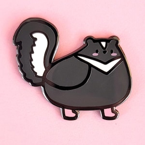 May include: A black and white skunk enamel pin with pink accents. The skunk has a white stripe down its back and a white chevron on its chest. The pin is set against a pink background.