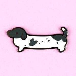 May include: Black and white enamel pin of a dachshund with black spots. The dachshund is facing left and has a black nose and eyes.