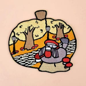 May include: Embroidered iron-on patch featuring a gray creature wearing a red hat and scarf, walking through a yellow and orange autumn scene with trees and mushrooms.