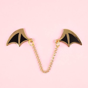 May include: A pair of gold-tone metal wings with black enamel accents connected by a gold chain. The wings are shaped like bat wings.