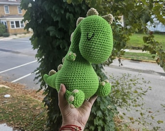 May include: A green crocheted dinosaur toy with a smiling face and small beige accents on its back, feet, and head. The handmade plushie is held up against a backdrop of green foliage and a residential street.