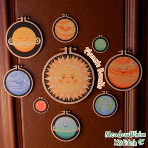 May include: A collection of embroidered planet designs in wooden hoops. The planets include a smiling sun, Jupiter, Earth, and others, all with smiling faces. The embroidery features orange, yellow, blue, and green colors. The text "Planets Bundle" is visible.