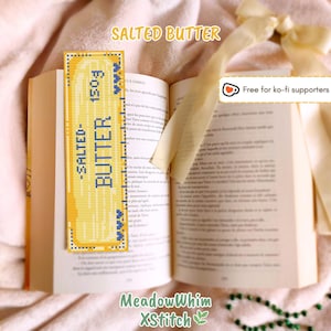 May include: A yellow and blue cross-stitch bookmark shaped like a butter stick, with the text "SALTED BUTTER 150g". The bookmark is placed in an open book. The text "SALTED BUTTER" is at the top of the image.