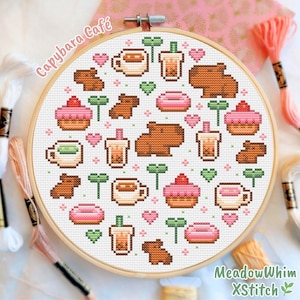 May include: A cross-stitch embroidery hoop featuring a repeating pattern of capybaras, coffee cups, bubble tea, cupcakes, donuts, and hearts. The text "Capybara Café" and "MeadowWhim XStitch" are visible. The color palette includes brown, pink, green, and white.