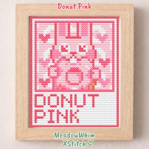 May include: A framed cross-stitch artwork featuring a pink donut design. The artwork includes the text "Donut Pink" at the top and bottom, with the artist's name and logo at the bottom. The frame is made of light-colored wood.