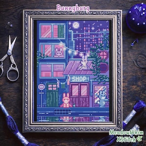 Peut inclure: Une œuvre d'art au point de croix dans un cadre argenté représente un paysage urbain nocturne avec une boutique, des bâtiments et des lapins. La palette de couleurs comprend des nuances de bleu, de violet et de rose. Le texte "Bunnyburg" et "SHOP" sont visibles.